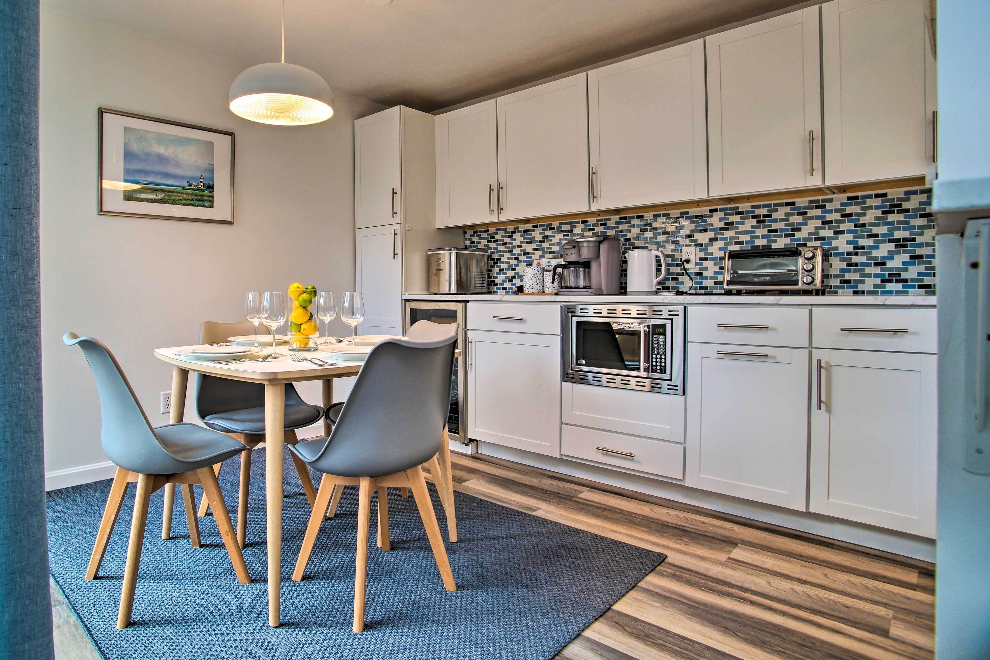 A modern kitchen with white cabinets, a microwave, a toaster, and a coffee maker. There is a dining table with chairs and a blue rug.