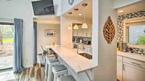 Une cuisine moderne dotée d’un comptoir en marbre, de tabourets de bar, d’un éclairage suspendu et d’une grande fenêtre offrant une vue sur l’extérieur.