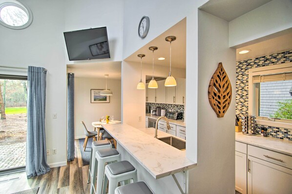 Una cocina moderna con encimera de mármol, taburetes de bar, iluminación colgante y un amplio ventanal con vistas al exterior.