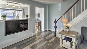 A modern living room with a large flat-screen TV, wooden flooring, a staircase with a lamp, and a view of the outside. - Pet-friendly Revere Getaway, Steps From the T! (Revere)