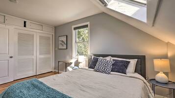 Quarto com claraboia, cama com almofadas estampadas, luminária de cabeceira e janela com vista para o verde.