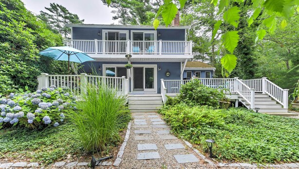 A two-story house with a front deck, a pathway leading to the entrance, and a garden with hydrangeas and shrubs. - Mashpee House w/ Wraparound Deck - Walk to Beach! (Mashpee)