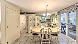 A dining area with a round table and chairs, a kitchen in the background, and a view through a glass door. - Mashpee House w/ Wraparound Deck - Walk to Beach! (Mashpee)