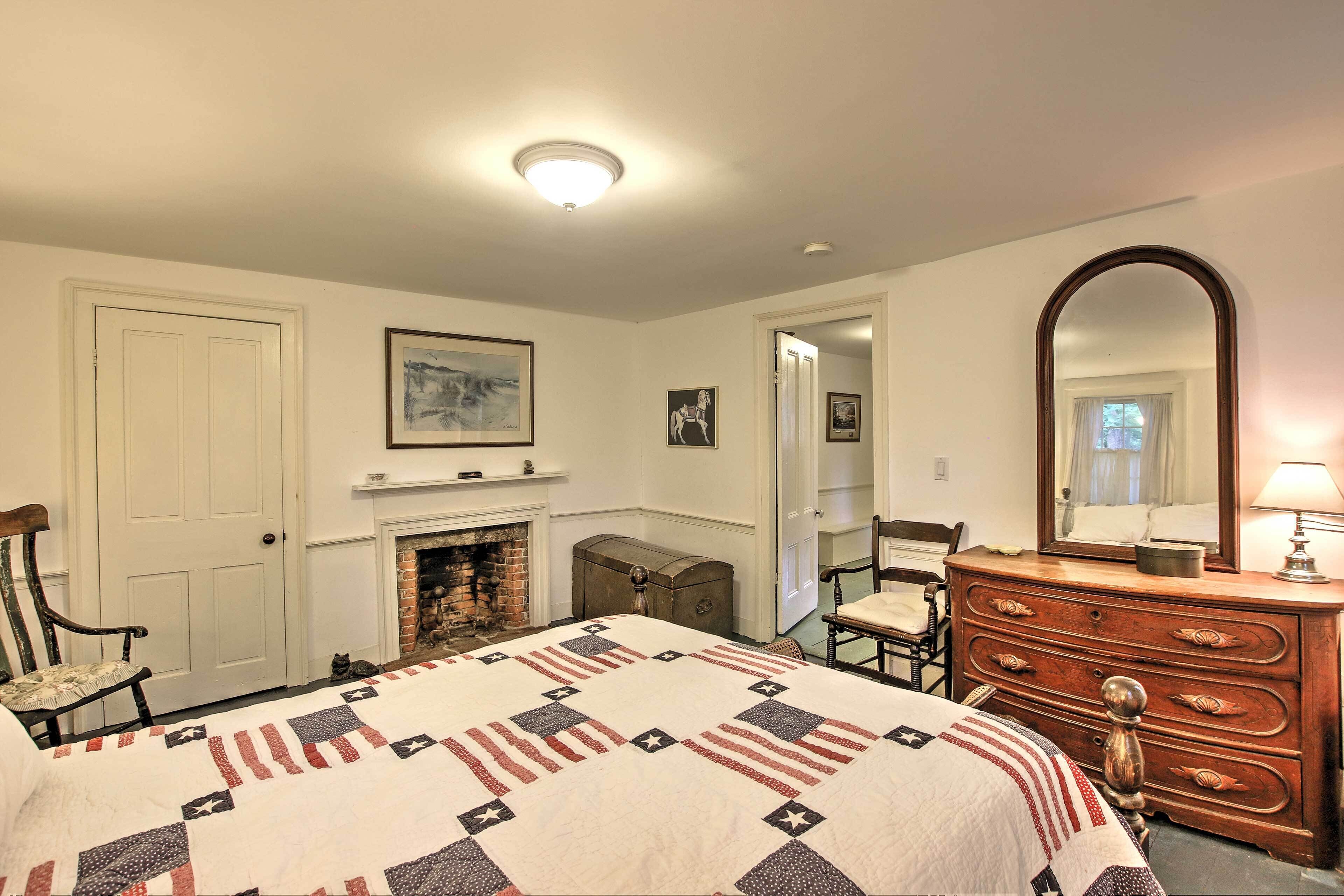 A bedroom with a bed, dresser, chair, fireplace, and a mirror.