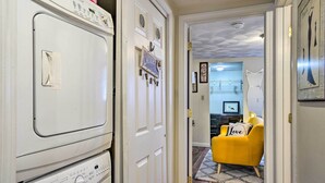 Une buanderie avec des sèche-linge superposés et une porte donnant sur un espace de vie où se trouvent un fauteuil jaune et un coussin orné du mot « Love ».