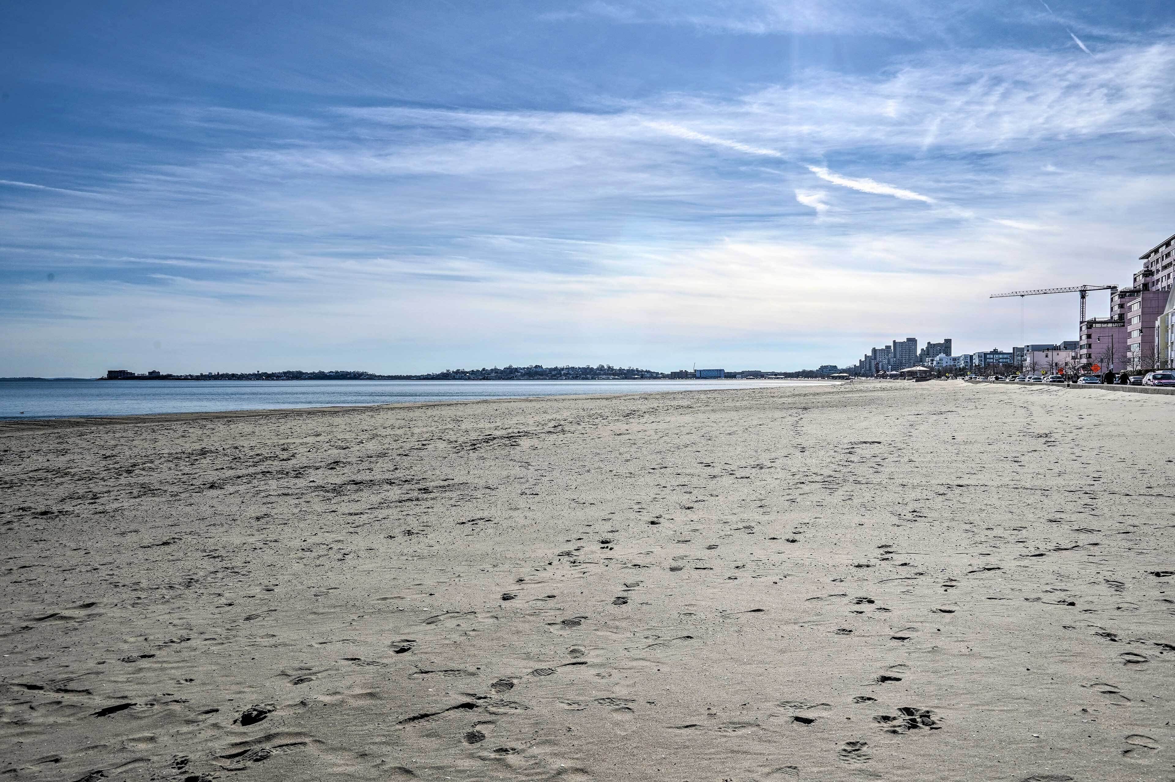 A wide sandy beach with footprints, a clear sky, and buildings in the background.