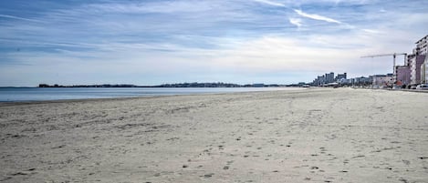 A wide sandy beach with footprints, a clear sky, and buildings in the background.