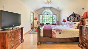 A bedroom with a large flat-screen TV, a bed with floral bedding, a chandelier, and a view of trees through a window. - Granby Home w/ Deck < 1/2 Mi to Aldrich Lake! (Granby)