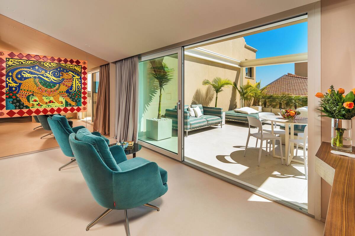 Una habitación de hotel moderna con sillas color turquesa, un amplio ventanal que da a un balcón con zona de comedor y un tapiz colorido en la pared.