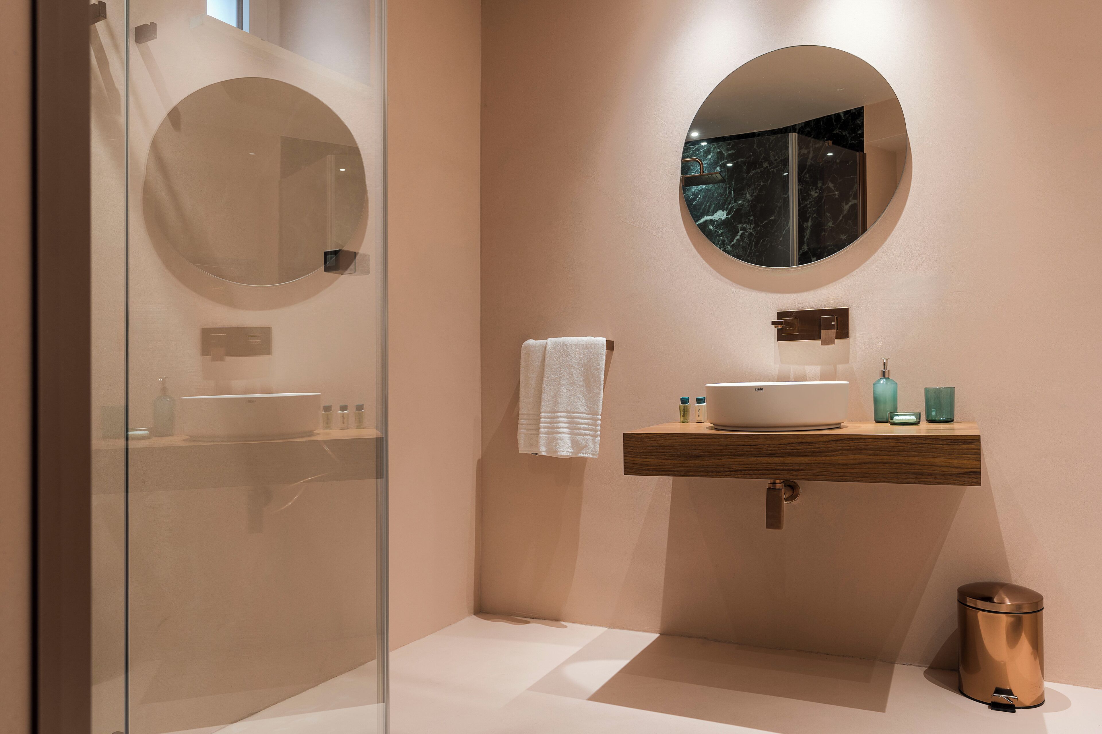 Une salle de bain moderne avec une douche à parois de verre, un miroir rond, un meuble-lavabo en bois et une poubelle.