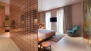 A modern hotel room with a large bed, a seating area with a round table, a blue armchair, a TV mounted on the wall, and a decorative wall mural. - Taormina Infinity Suites (Taormina)