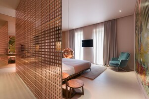 A modern hotel room with a large bed, a seating area with a round table, a blue armchair, a TV mounted on the wall, and a decorative wall mural.