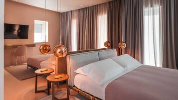 Quarto de hotel moderno com uma cama grande, um espaço de estar com uma poltrona e uma mesa pequena, e uma televisão fixada na parede.