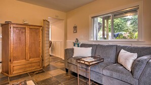 Ein Wohnzimmer mit grauem Sofa, Holzschrank und Fenster mit Blick auf Bäume.