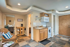 A compact kitchen with a built-in oven, a microwave, and a small island with a sink. There is a wicker chair and a small table with magazines. - Brookings Vacation Rental Studio - Walk to Town! (Brookings)