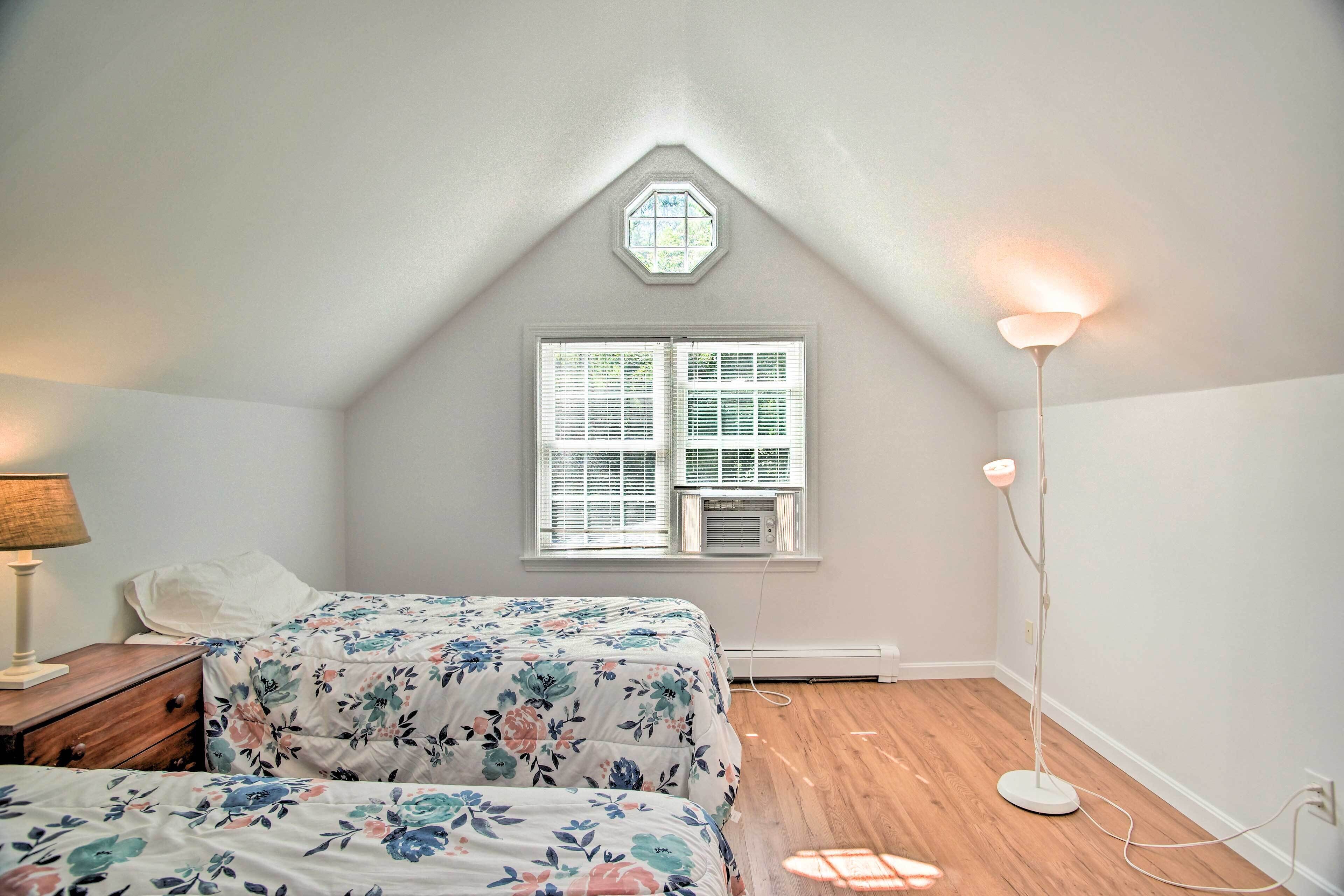 A bedroom with a sloped ceiling, a skylight, a window with blinds, a bedside table with a lamp, and a floor lamp.