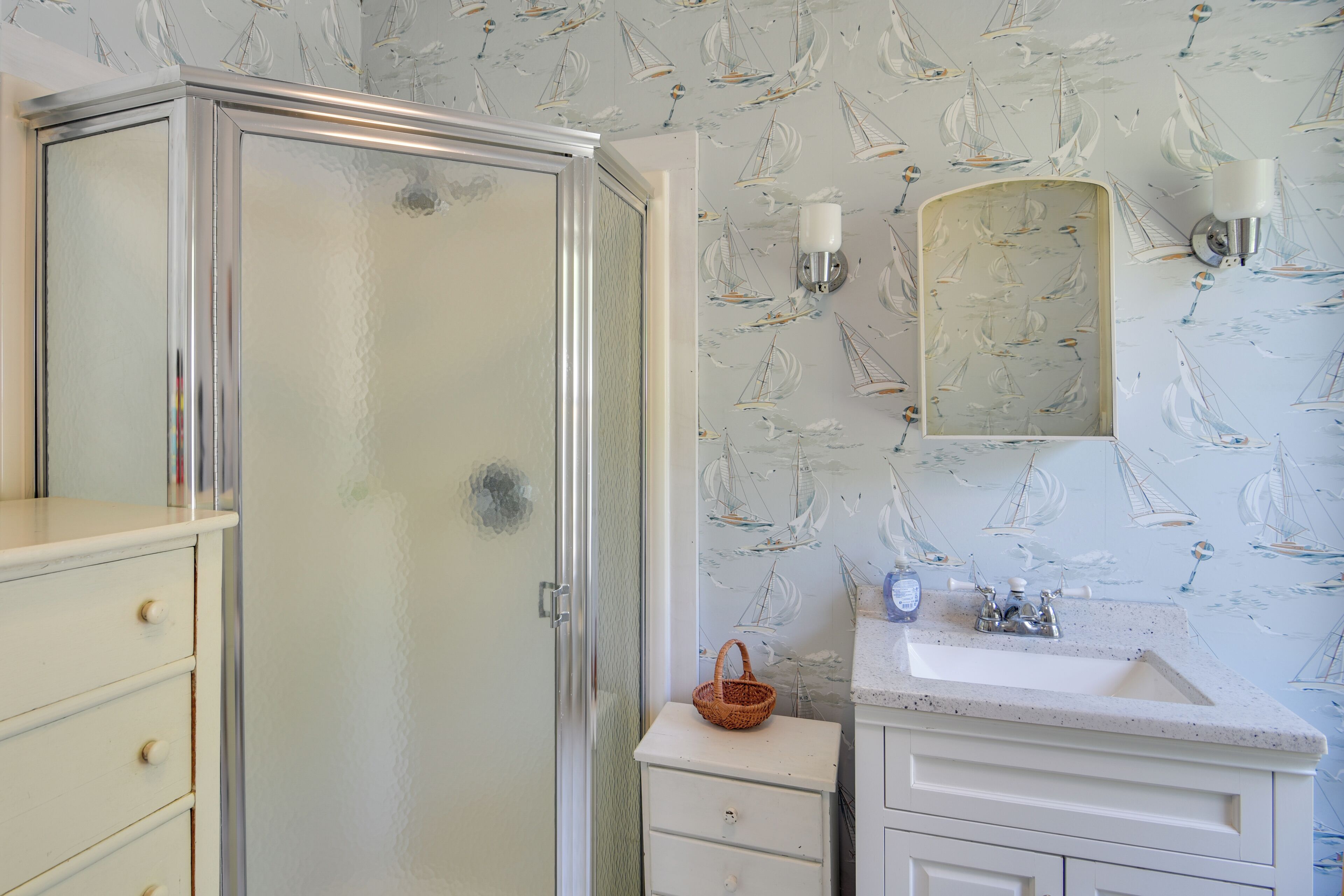 A bathroom with a glass shower enclosure, a sink with a mirror, and a patterned wallpaper.