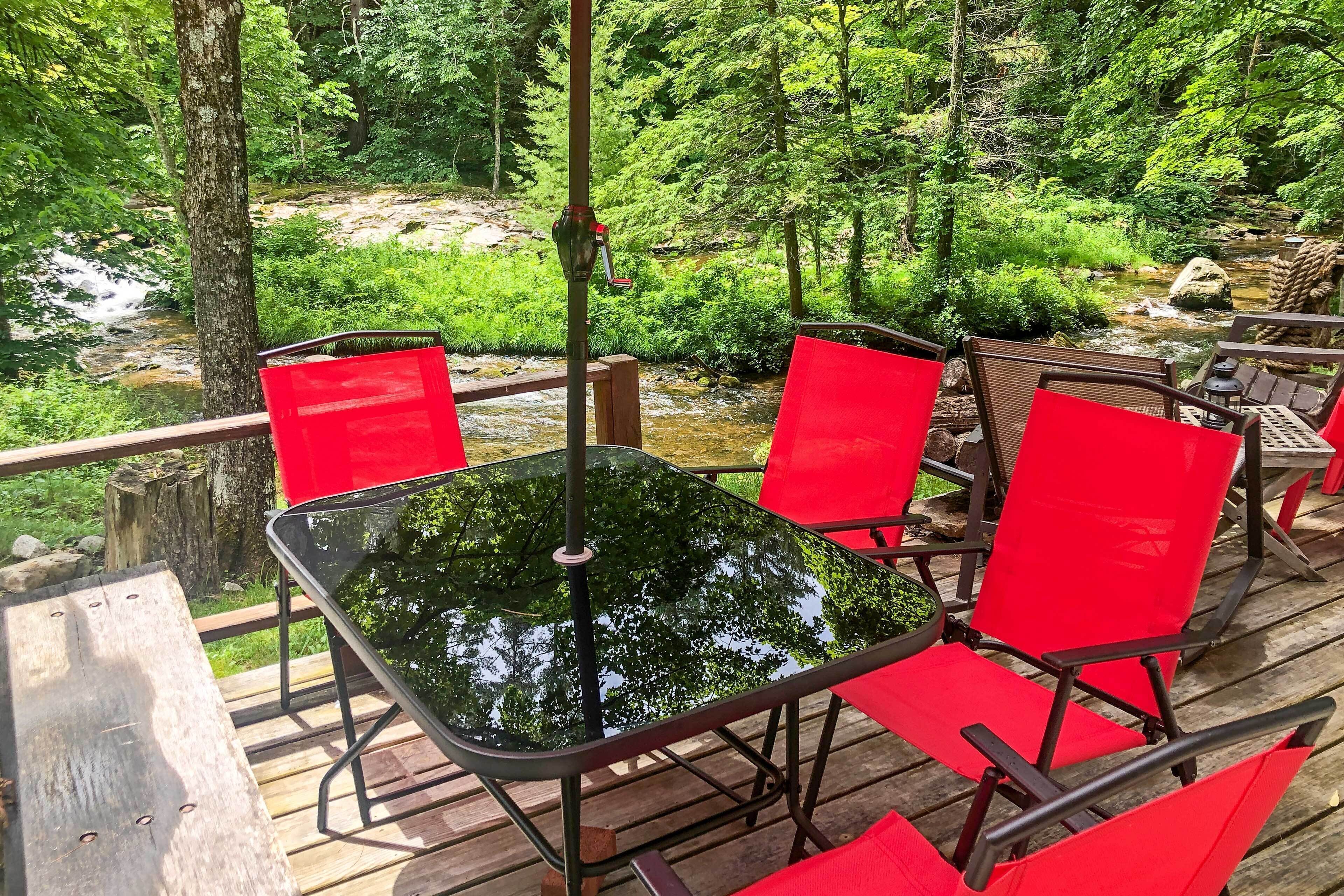A wooden deck with a glass table and red chairs overlooking a stream and forest.