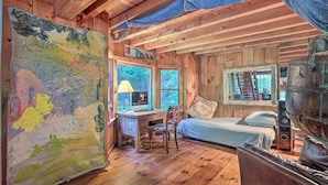 A rustic cabin interior with a wooden desk, a bed, and a large abstract painting. - Serene Riverfront Stay! 'the Mill River Cabin (Mill River)