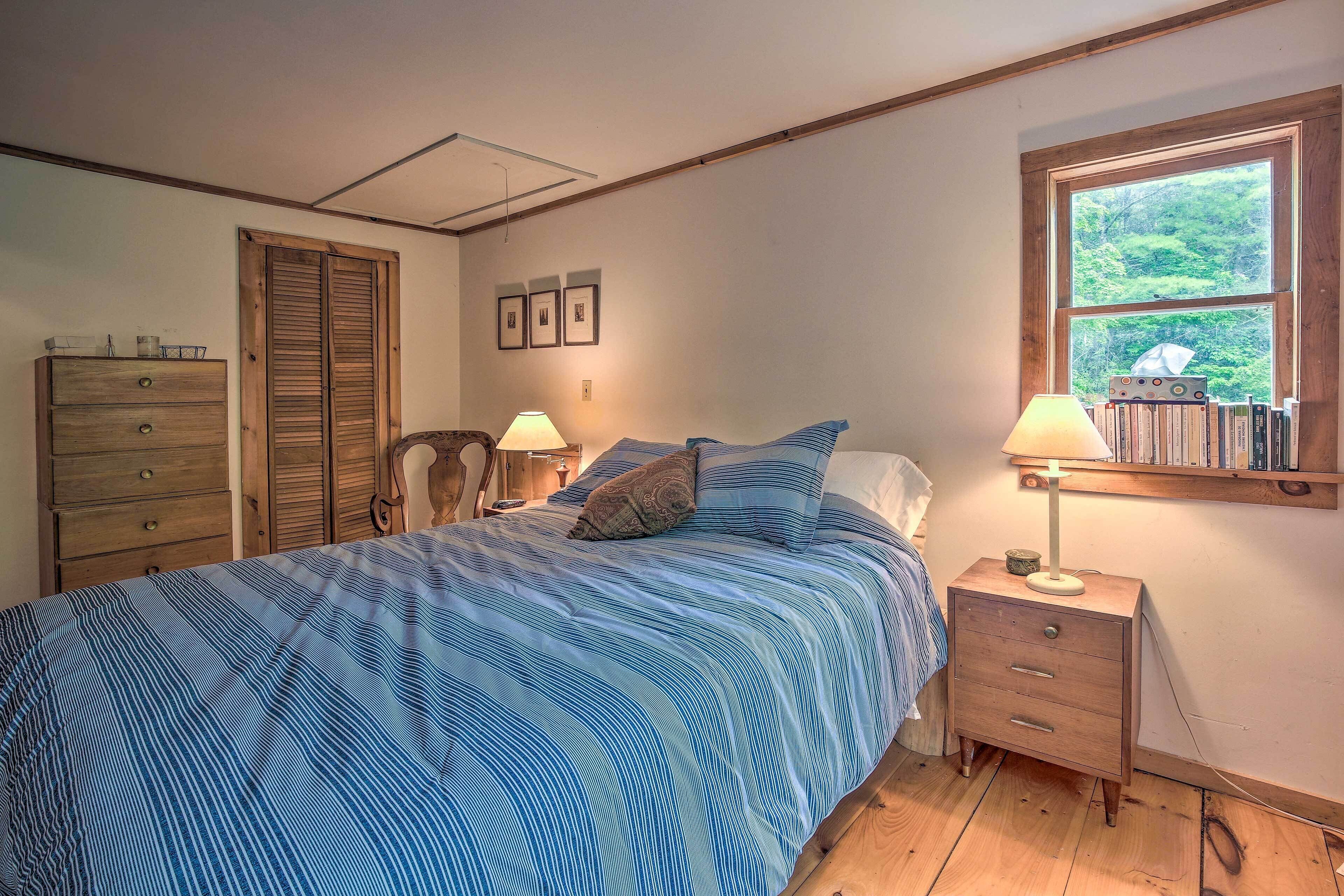 A bedroom with a bed, wooden nightstand, dresser, and a window with a view of greenery.