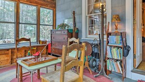 A rustic cabin interior with wooden walls, a large window, a wooden table with chess pieces, a bookshelf, and a lamp. - East Otis Reservoir Cabin w/ Porch - Walk to Lake! (Otis)
