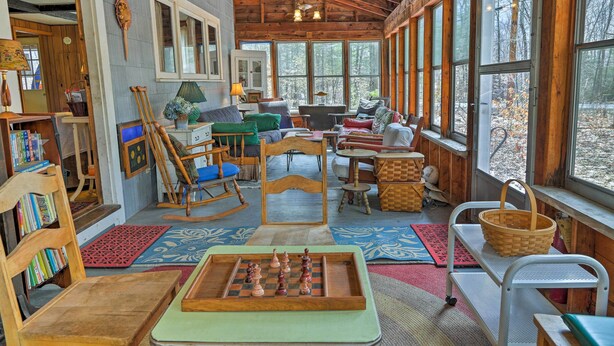 A cozy cabin interior with a wooden ceiling, large windows, a chessboard, and a bookshelf. - East Otis Reservoir Cabin w/ Porch - Walk to Lake! (Otis)