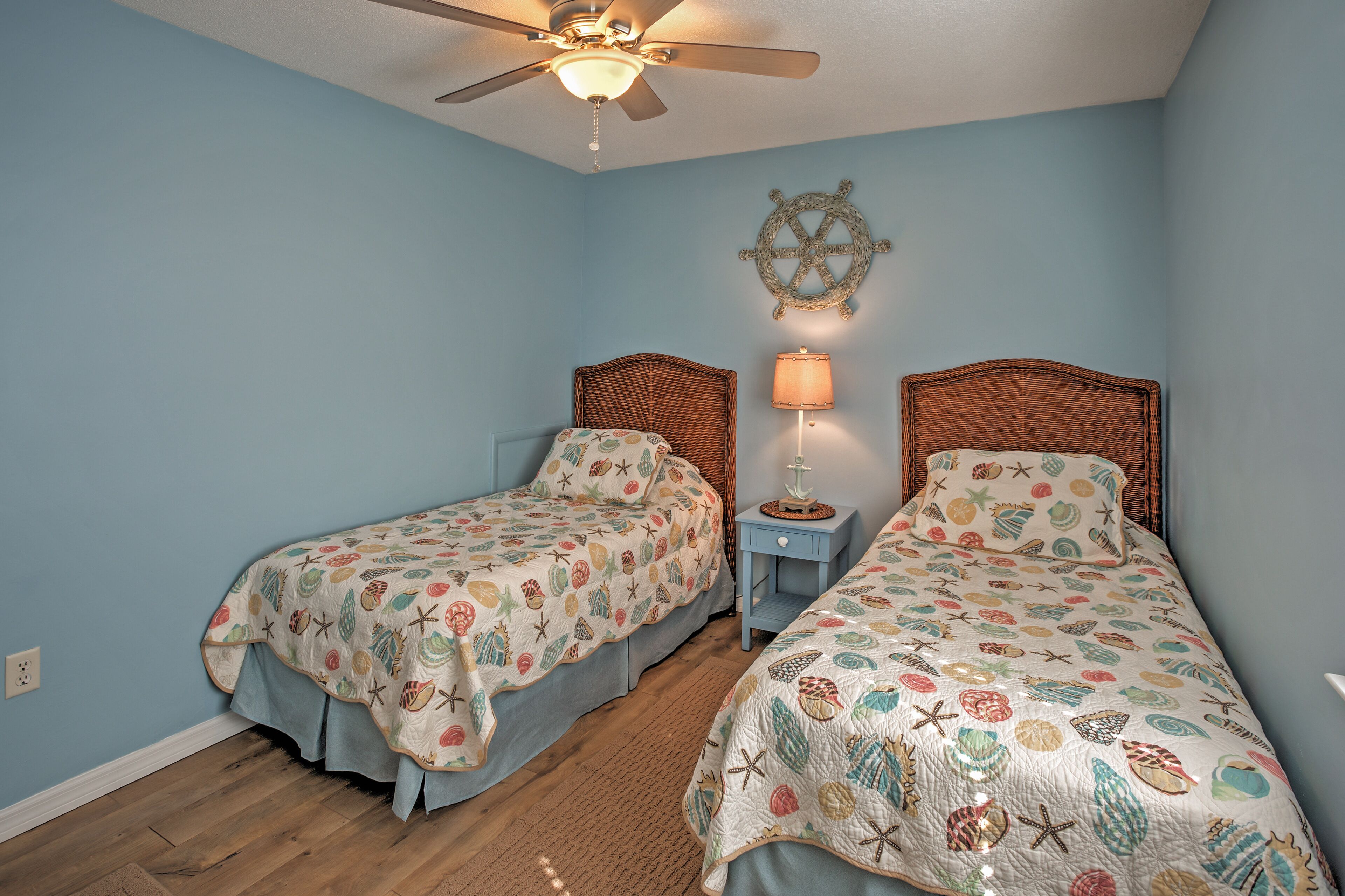 A bedroom with two beds, a ceiling fan, a ship's wheel decoration, and a nightstand with a lamp.
