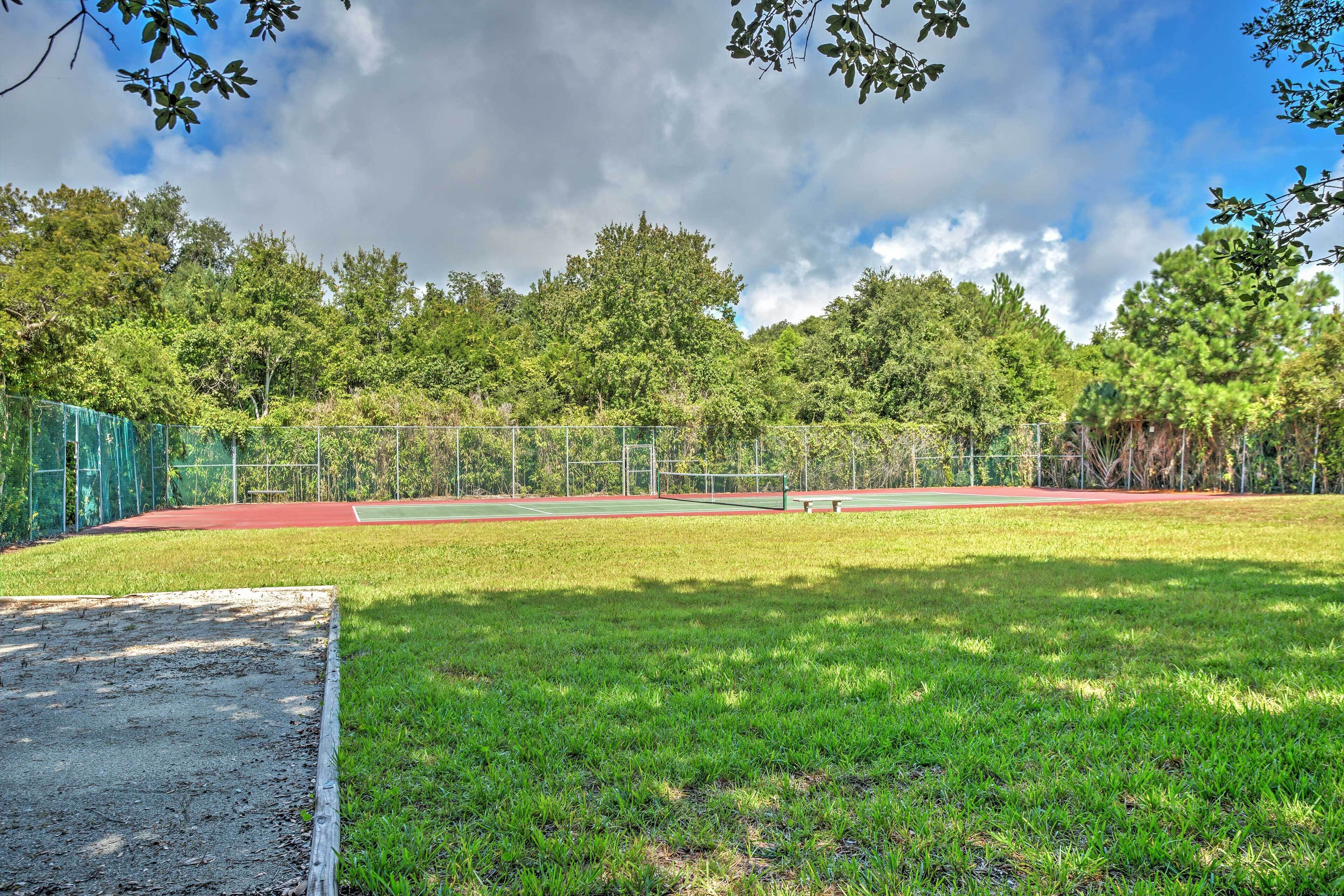 A tennis court with a green fence, surrounded by trees and a cloudy sky.