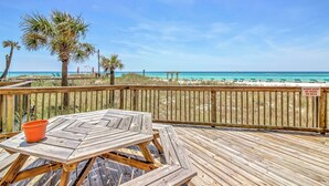 A wooden deck with a table and chairs, overlooking a beach with palm trees and a clear blue sky. - Bright PCB Condo w/ Gulf Views & Beach Access! (Panama City Beach)