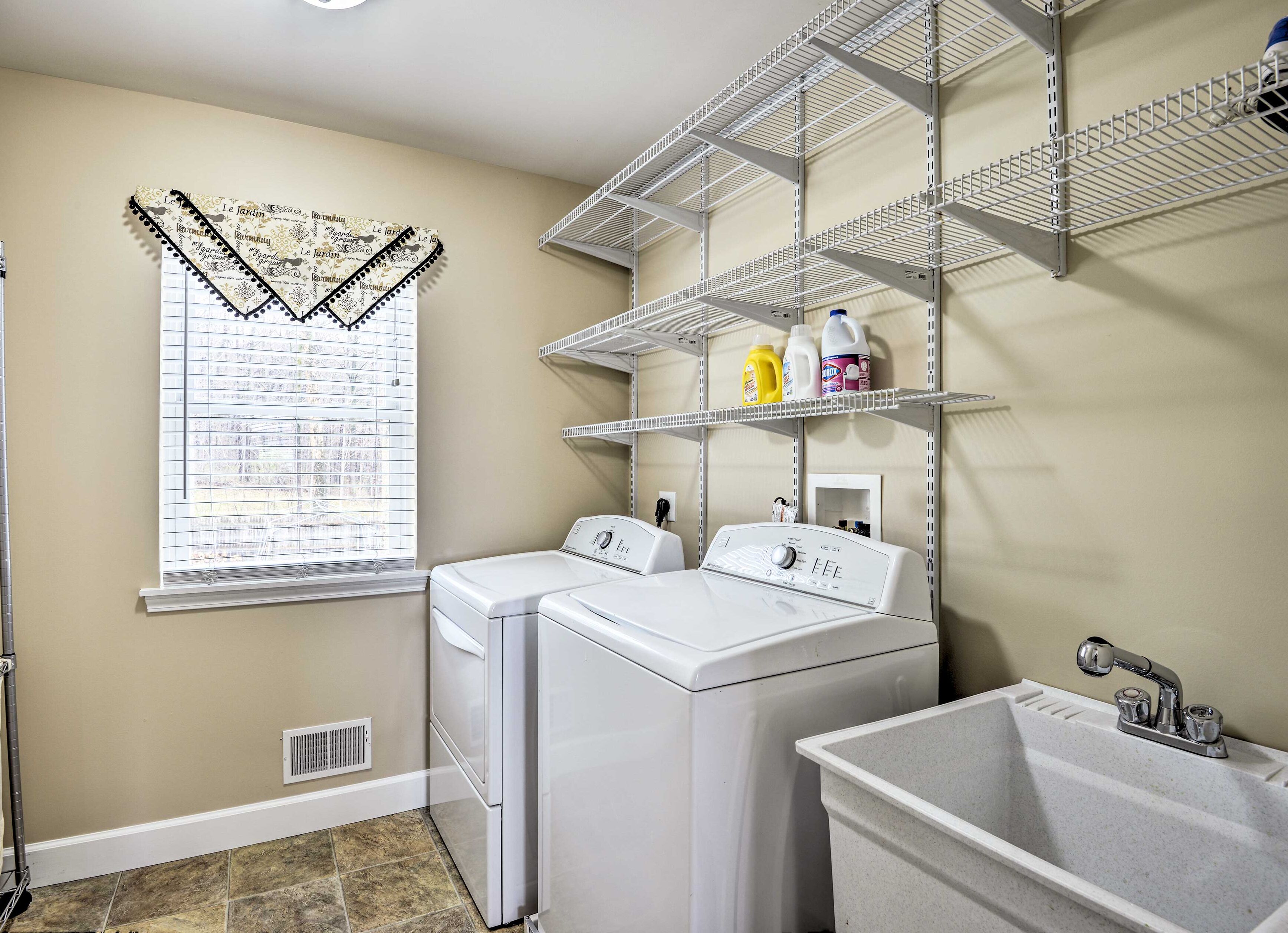 A laundry room with two white front-loading washers, a metal shelving unit, a sink, and a window with decorative blinds.