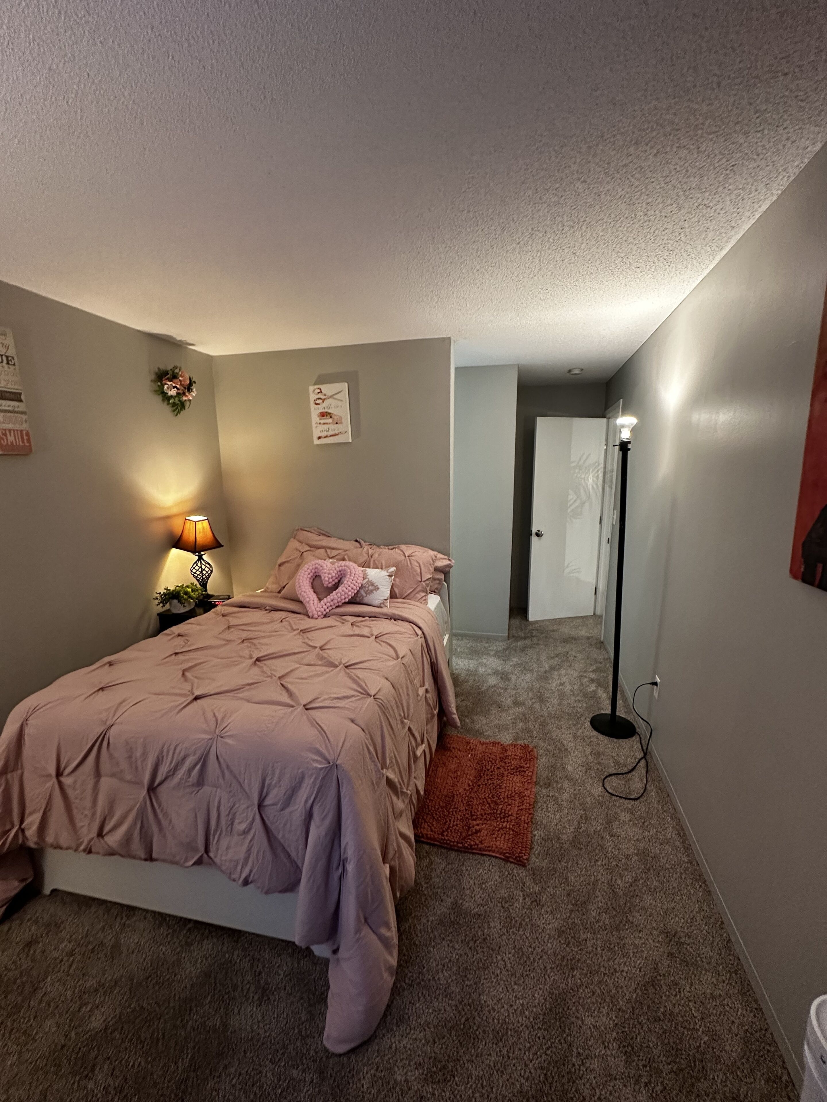 Un dormitorio con una cama, una mesita de noche con lámpara, una puerta y una lámpara de pie.