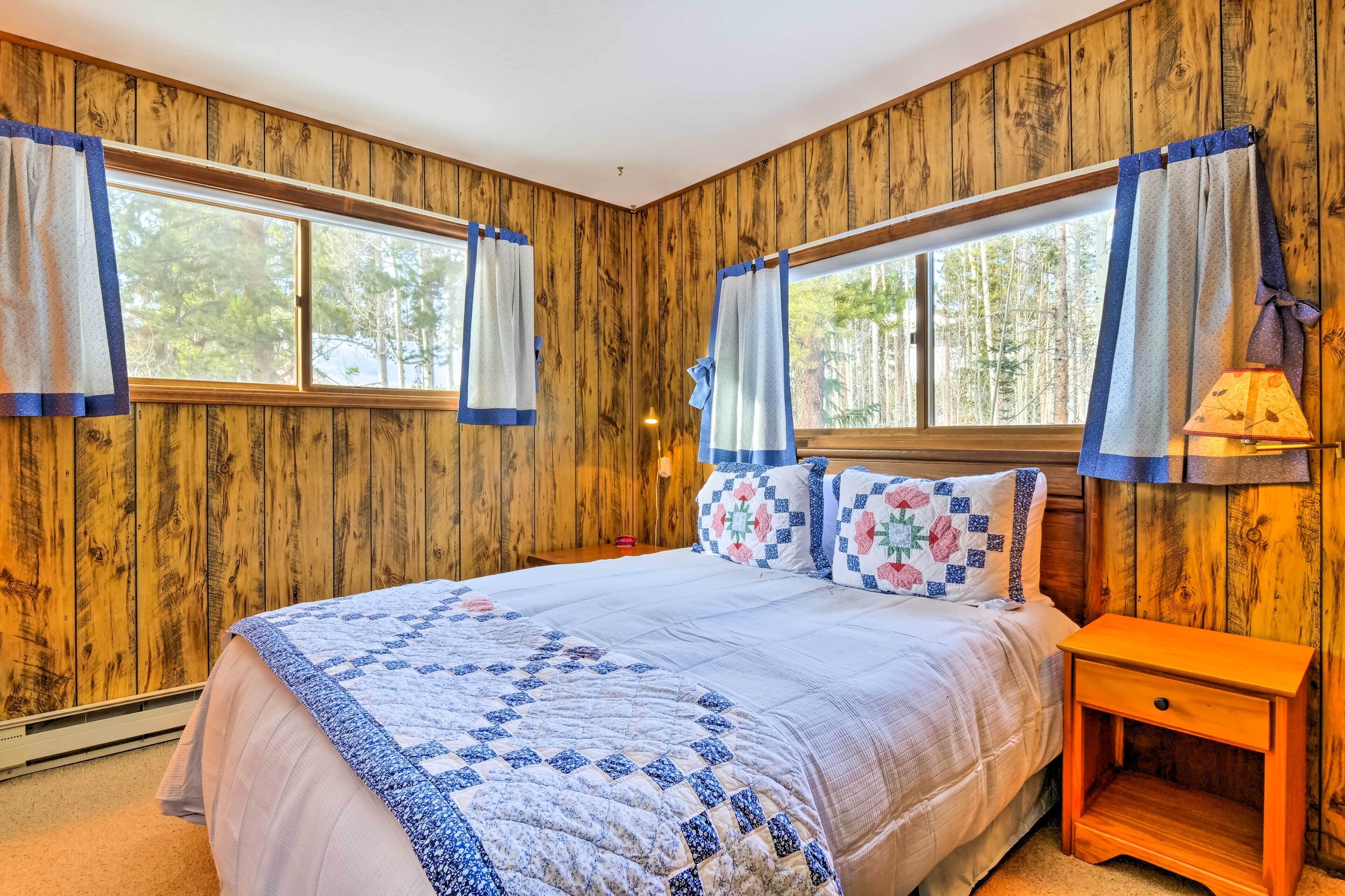 Une chambre avec des murs en bois, un grand lit recouvert d’une courtepointe, deux fenêtres avec des rideaux et une table de chevet avec une lampe.