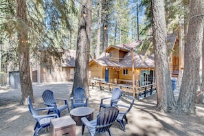 A wooden cabin with a deck, surrounded by tall trees. There are Adirondack chairs and a table with a box.