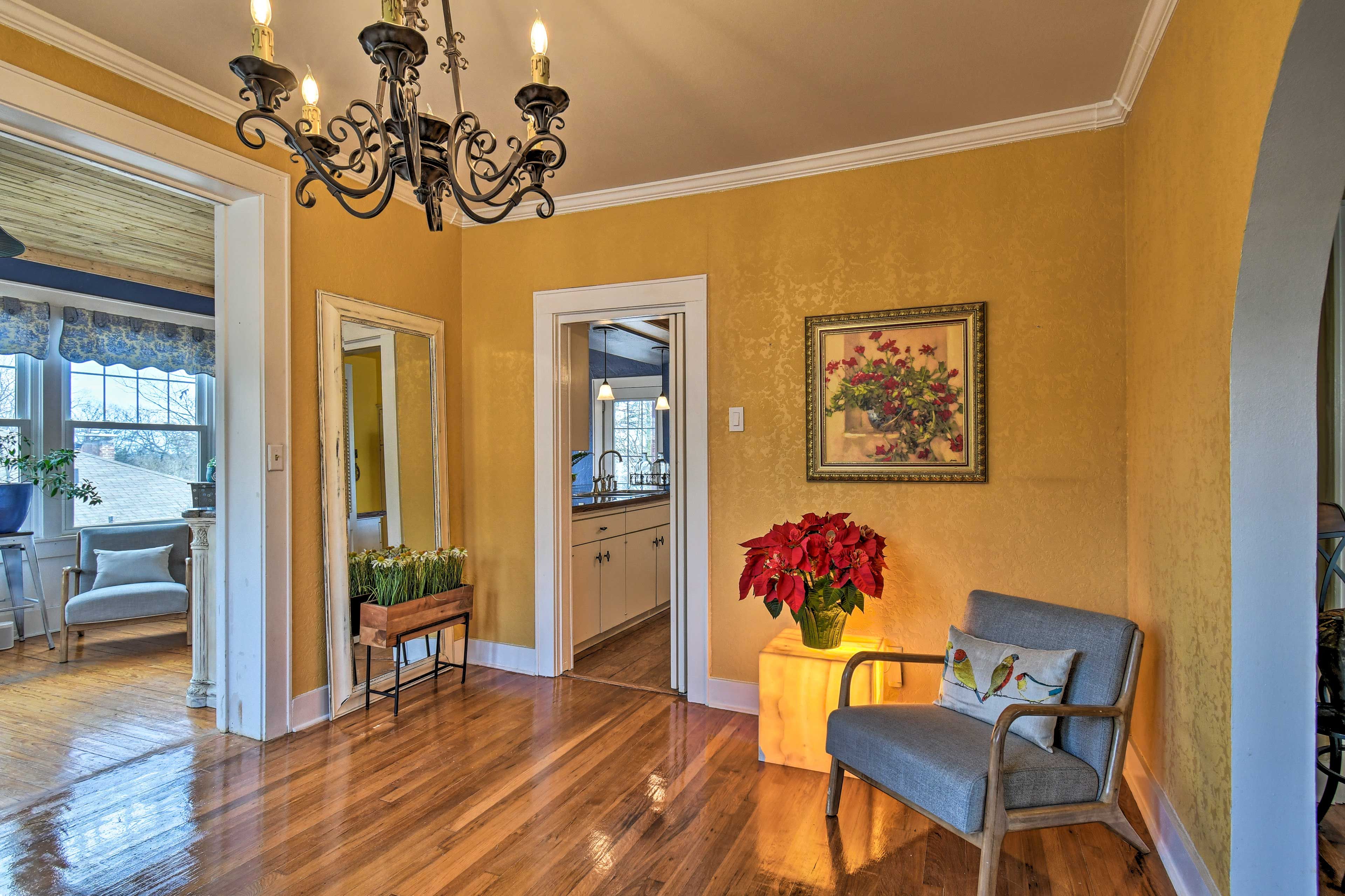 Un ampio soggiorno con parquet, un lampadario, una panca, un tavolo con un vaso di fiori e una cucina visibile attraverso un'apertura.
