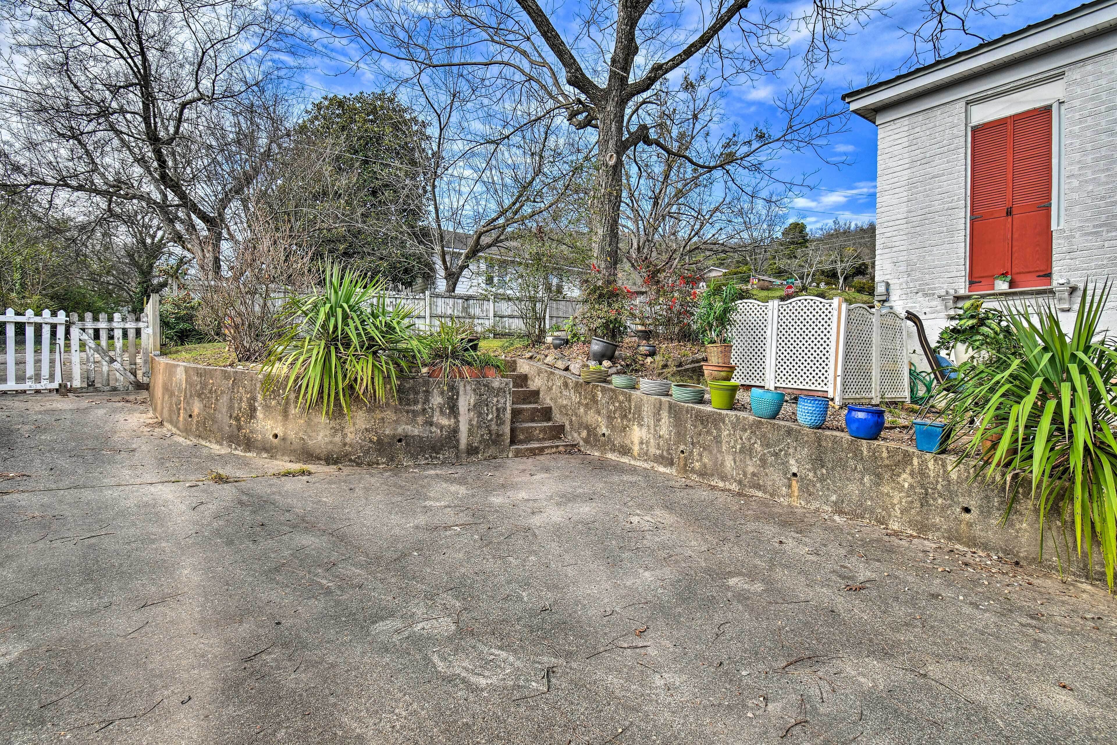 A concrete driveway leading to a house with a white picket fence, a red shuttered window, and a variety of potted plants.