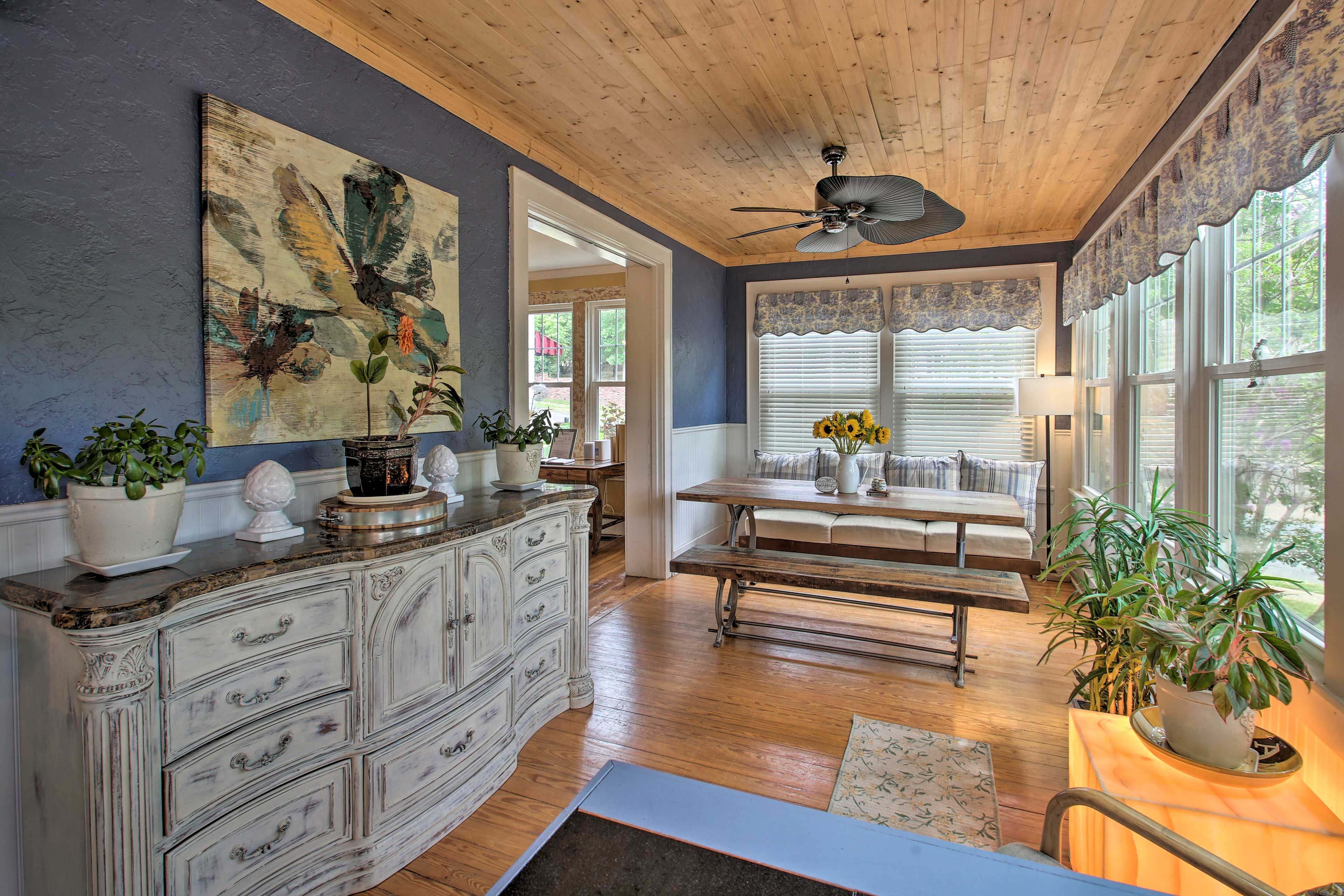 A room with a wooden ceiling, a ceiling fan, a large window with blinds, a dining table with chairs, and a white cabinet with drawers.