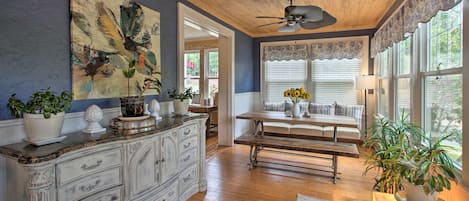A room with a wooden ceiling, a ceiling fan, a large window with blinds, a dining table with chairs, and a white cabinet with drawers.