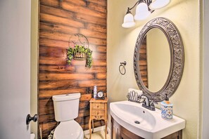 A bathroom with a wooden wall, a toilet, a sink with a mirror, and a small shelf with a plant and a clock. - Albuquerque Vacation Home Rental w/ Hot Tub! (Albuquerque)
