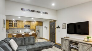 A modern living room with a gray sectional sofa, wooden cabinets, a flat-screen TV, and a kitchen in the background. - Cozy Avon Condo w/ Grill: Walk to Ski Shuttle (Avon)