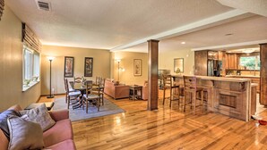 A spacious living room with a wooden floor, a sofa, a dining area with chairs, and a kitchen with wooden cabinets and a bar.