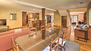 A spacious living area with a large glass-top dining table, wooden chairs, a sofa, and a kitchen in the background.