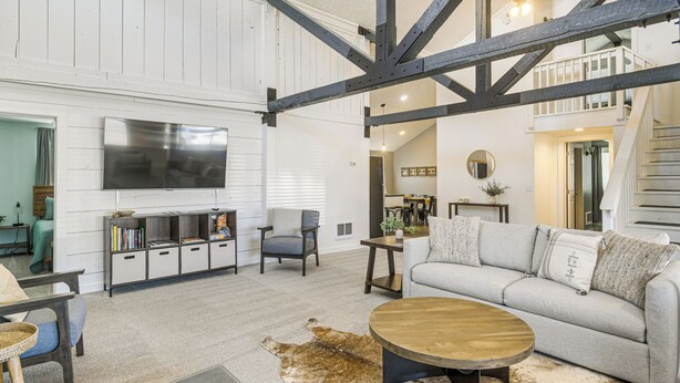 A spacious living room with a high ceiling, exposed beams, a grey sofa, a wooden coffee table, and a mounted TV. - Centrally Located Pinedale Vacation Rental! (Pinedale)