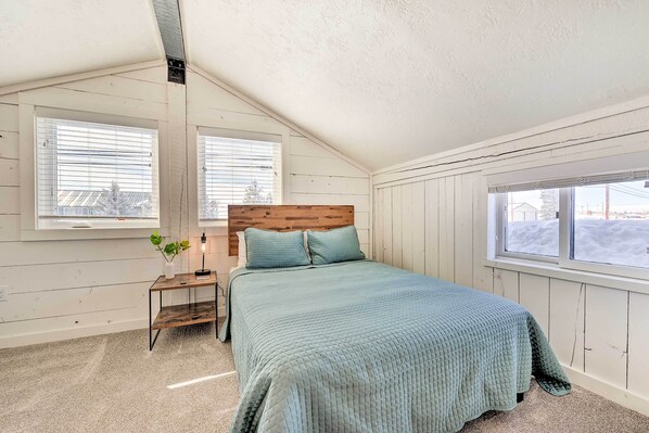 Un dormitorio con techo inclinado, paredes de madera, una cama con colcha verde, una mesita de noche con una planta y una ventana con persianas.