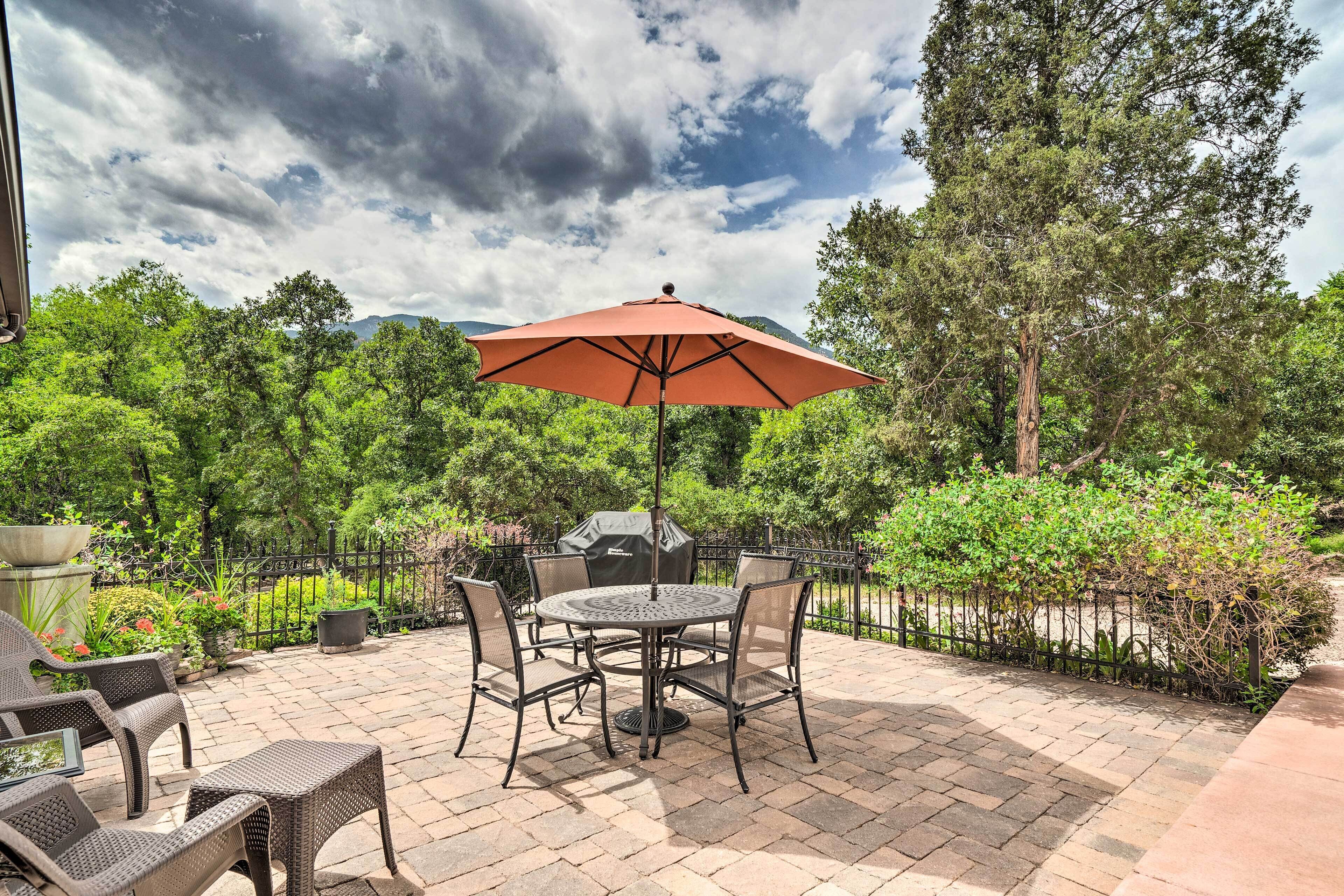 A patio with a round table and chairs, a red umbrella, and a grill.