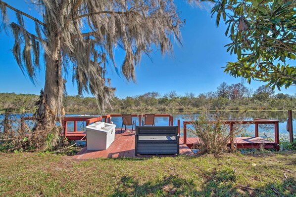 A wooden deck with a bench and chairs overlooking a body of water, surrounded by trees and a clear sky.
