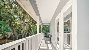 A covered porch with a white railing, a small seating area with a chair and a table, and a view of a wooded area.