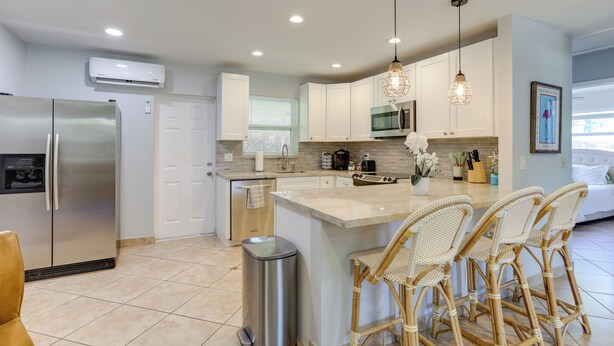 A modern kitchen with a stainless steel refrigerator, a built-in oven, a microwave, and a coffee maker. There are wicker chairs around a central island, and a trash can is visible. - 2 Mi to Swim & Shop: Updated North Palm Beach Home (Juno Beach)