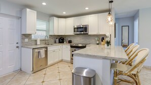 A modern kitchen with white cabinets, stainless steel appliances, a marble island, wicker chairs, and a trash can. - 2 Mi to Swim & Shop: Updated North Palm Beach Home (Juno Beach)