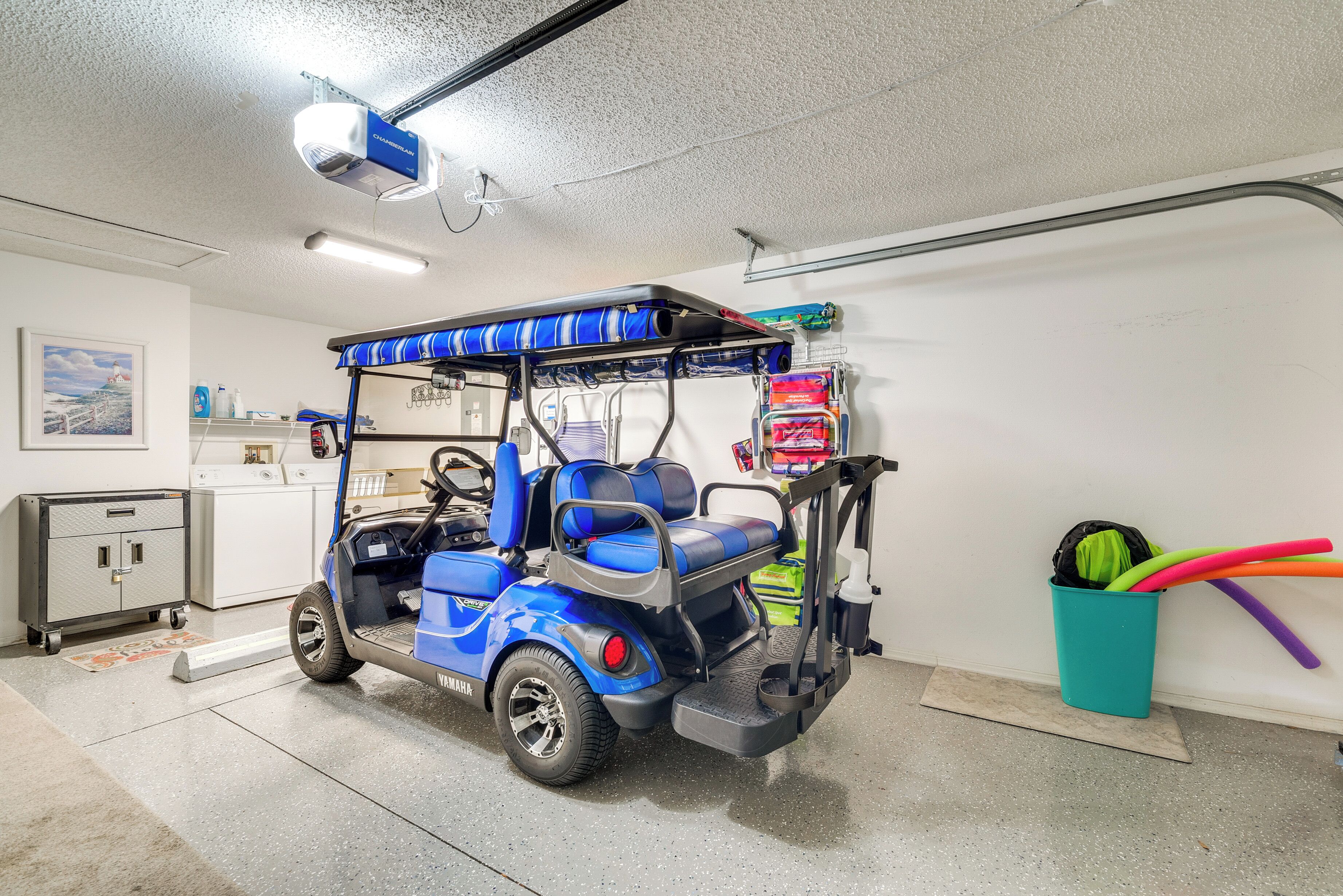 Ein blauer Golfwagen steht in einer Garage mit Deckenleuchte, einer Wandablage und einem Mülleimer.