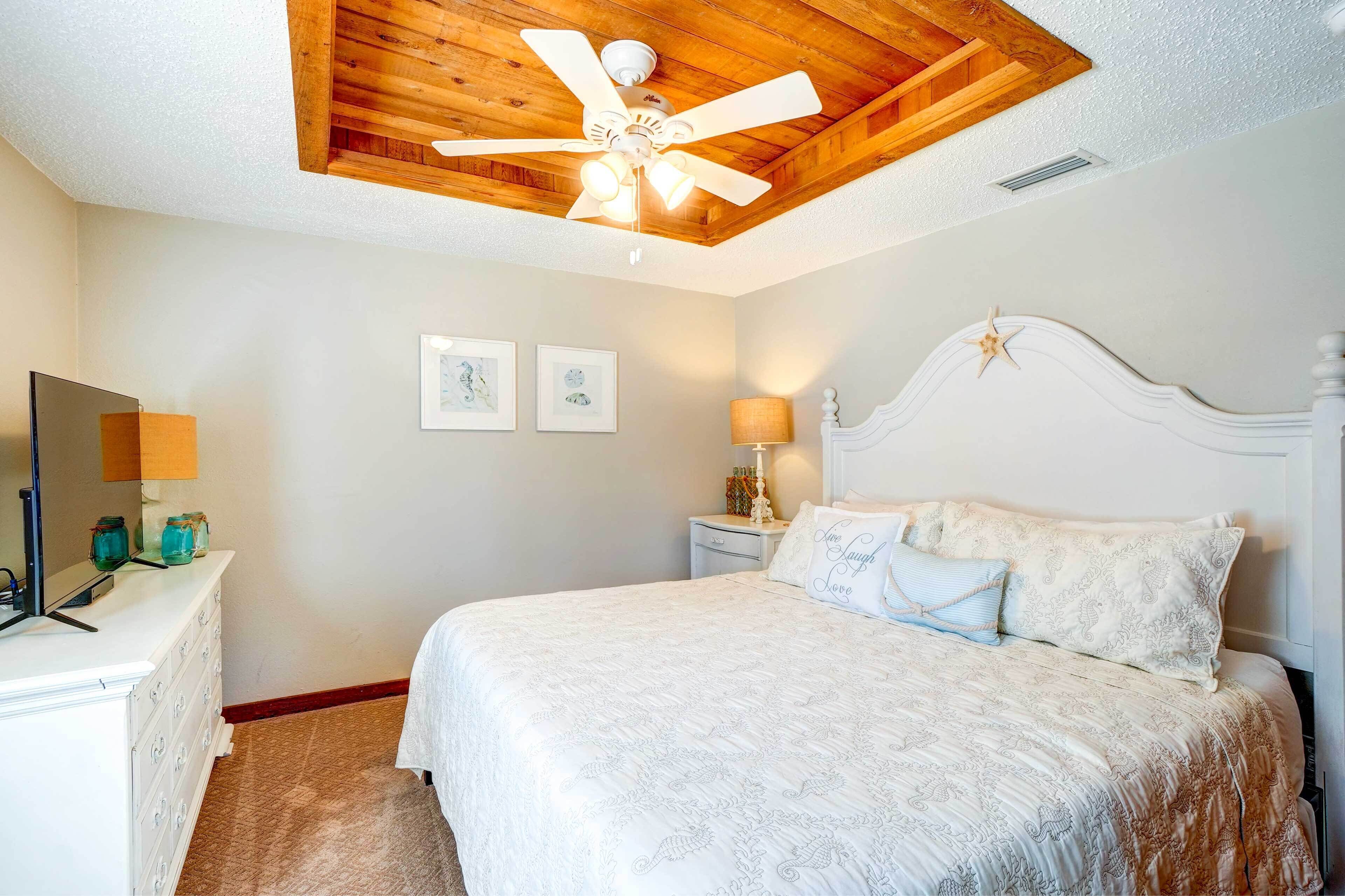 Un dormitorio con techo de madera, una cama con ropa de cama estampada, una mesita de noche con lámpara y cuadros en la pared.
